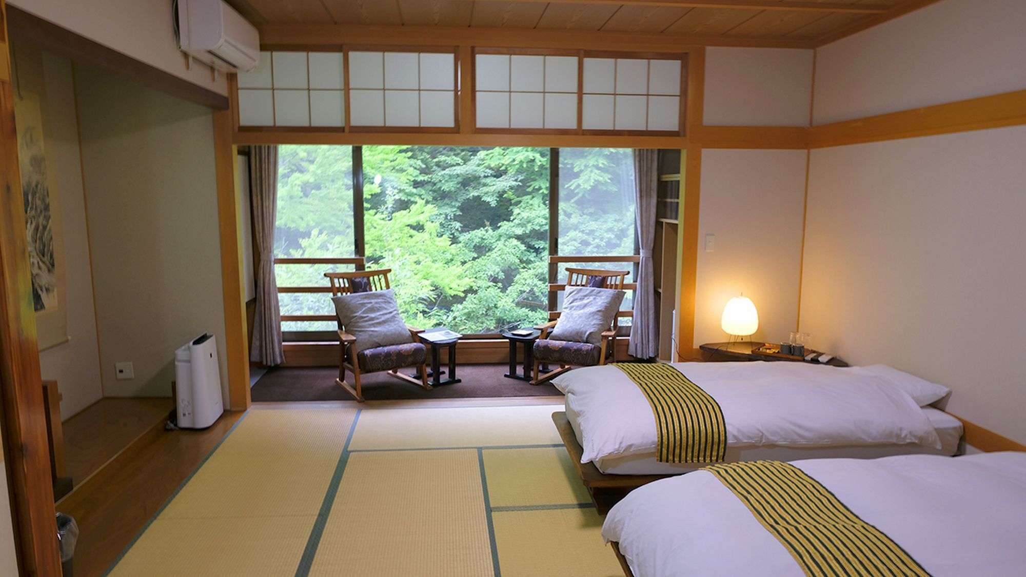 Een traditionele Japanse kamer met tatami vloerbedekking, een bed met gestreept beddegoed, twee schommelstoelen, een kleine tafel en een uitzicht op weelderige begroeiing door grote ramen.