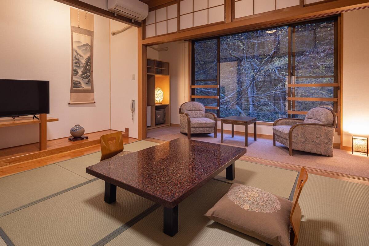 Een traditionele Japanse kamer met tatami vloeren, een lage tafel, stoelen, een televisie en een groot raam met uitzicht op bomen.