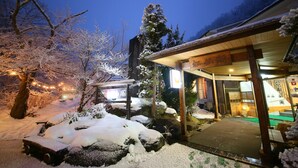 Un área exterior cubierta de nieve con la entrada de un edificio de madera, un letrero con caracteres japoneses y un camino de piedra que conduce a la entrada.
