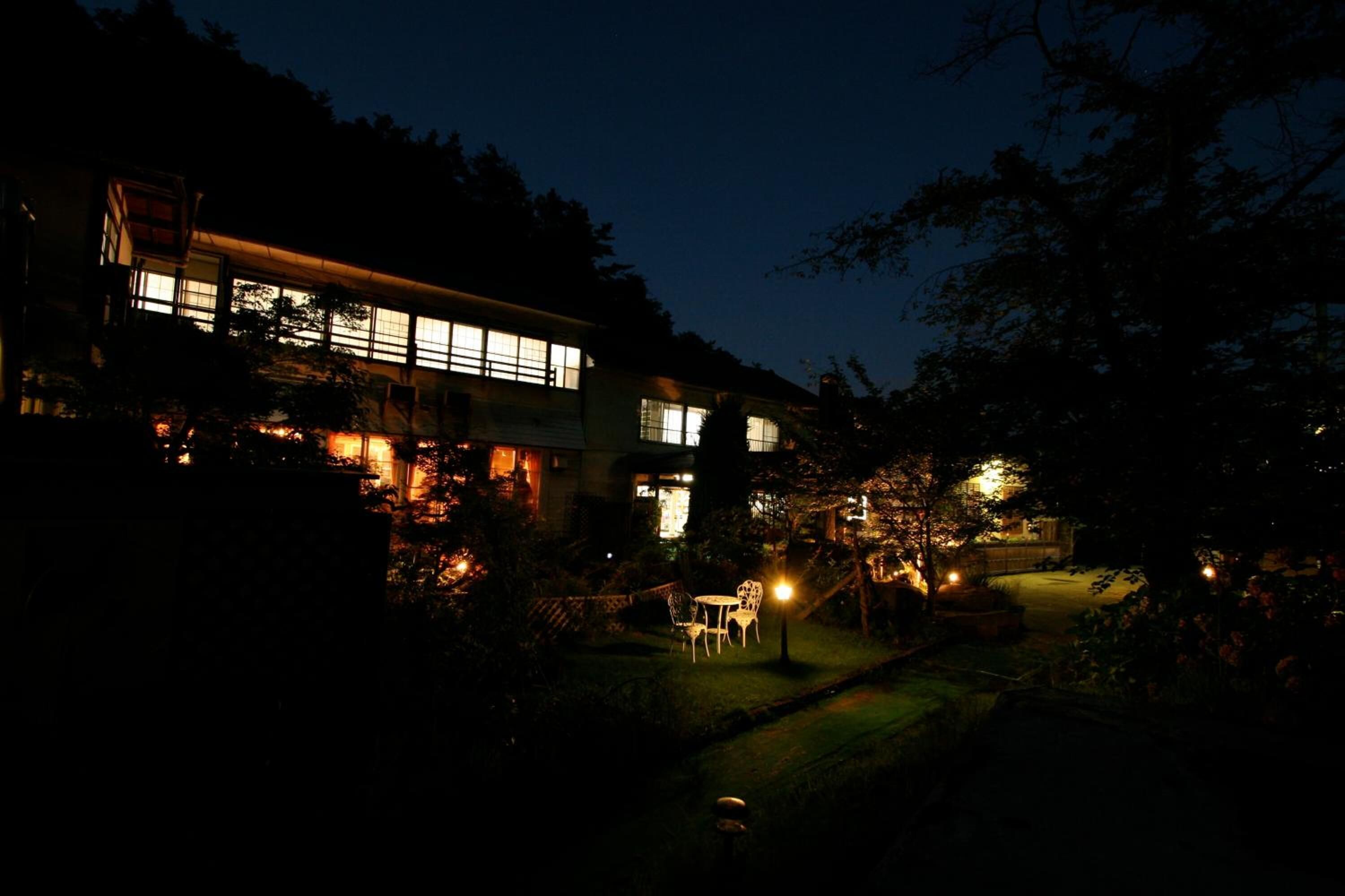 A two-story building with illuminated windows and a well-lit outdoor area with a table and chairs.