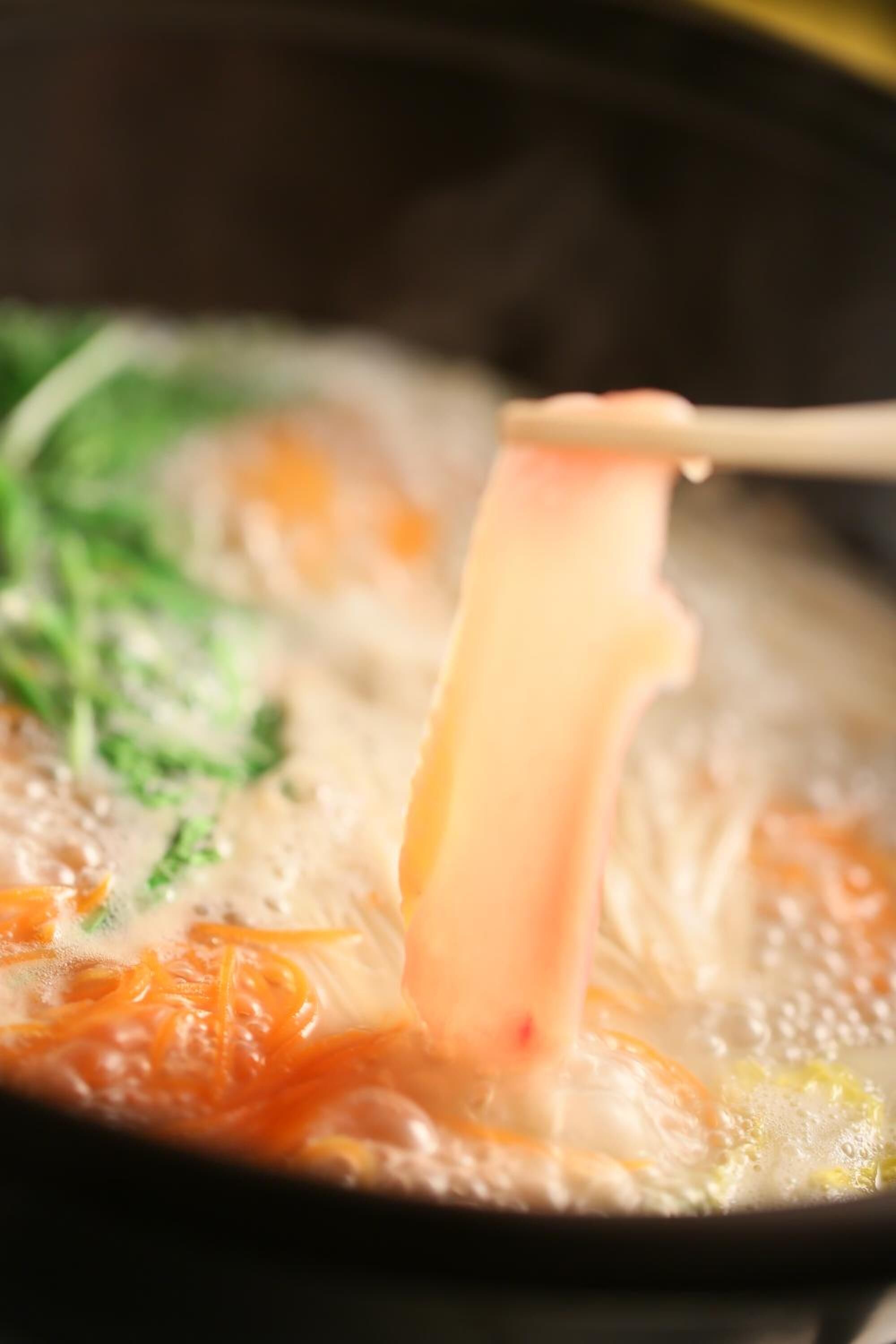 A close-up of a noodle dish being lifted with chopsticks, featuring a clear broth and visible vegetables.