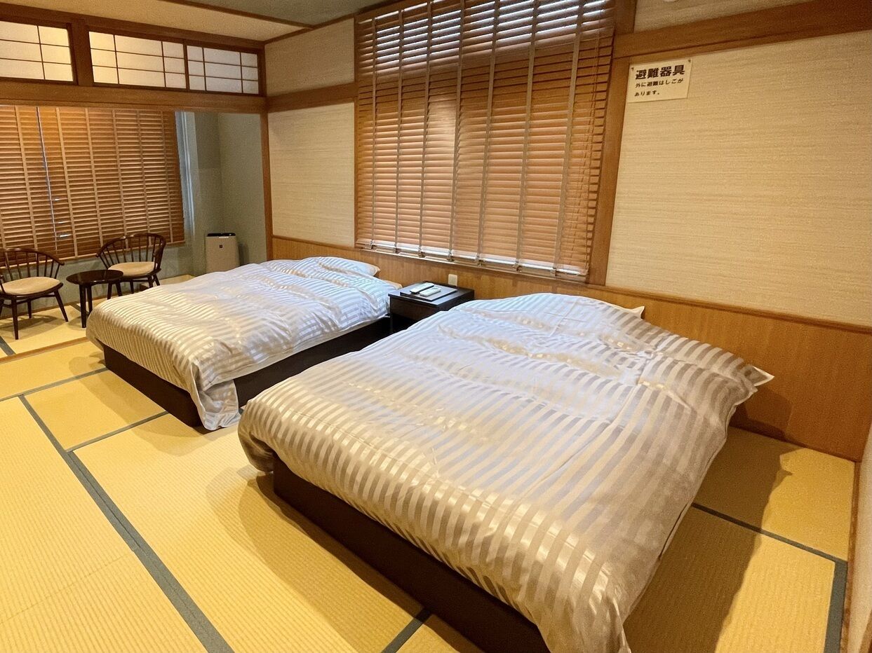 A traditional Japanese-style room with tatami flooring, two beds with striped bedding, a small table, and a chair.