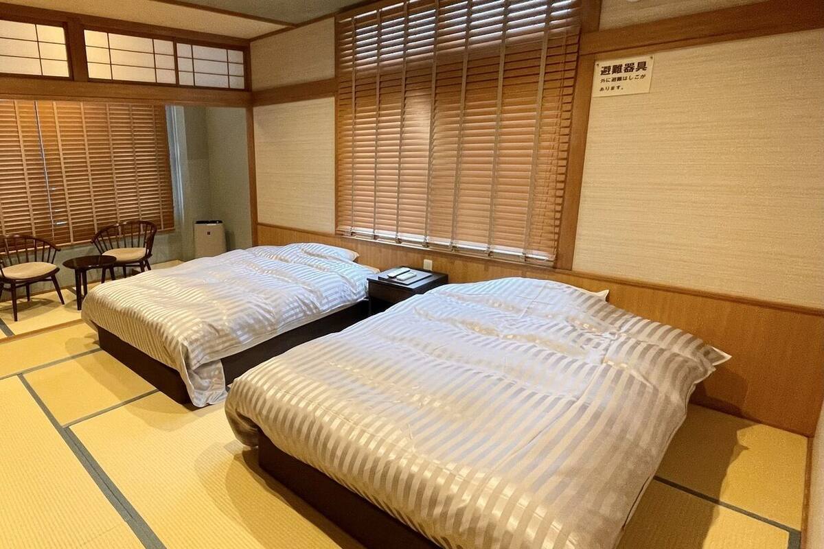 Ein traditioneller japanischer Raum mit Tatami-Böden, zwei Betten mit gestreifter Bettwäsche, einem kleinen Tisch und einem Stuhl.