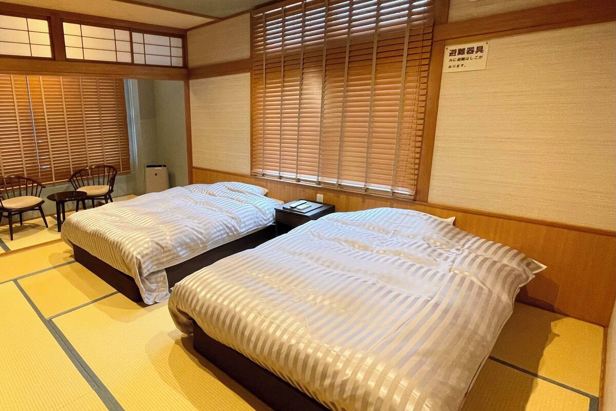 Ein traditioneller japanischer Raum mit Tatami-Boden, zwei Betten mit gestreifter Bettwäsche, einem kleinen Tisch, Stühlen und verschiebbaren Shoji-Türen.