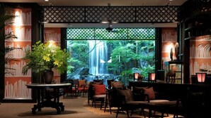 A cozy indoor seating area with a large window overlooking a waterfall, surrounded by bookshelves and decorative plants. - Konaya Hotel (Kofu)