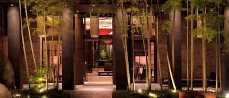 Un sendero bordeado por altos árboles e iluminado por una luz tenue, que conduce a un edificio con entrada iluminada en rojo.