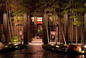 Un sendero bordeado por altos árboles e iluminado por una luz tenue, que conduce a un edificio con entrada iluminada en rojo.