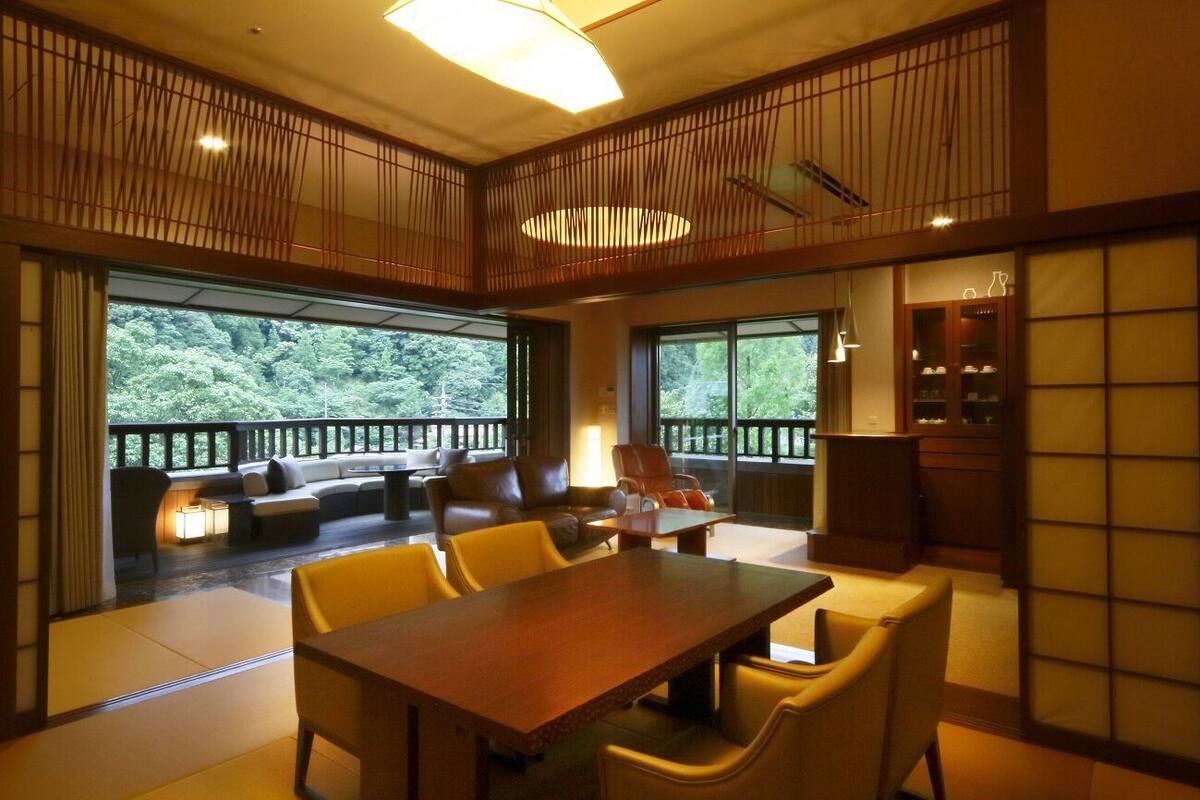 A spacious room with a wooden dining table, yellow chairs, a sofa, and a balcony with a view of greenery.