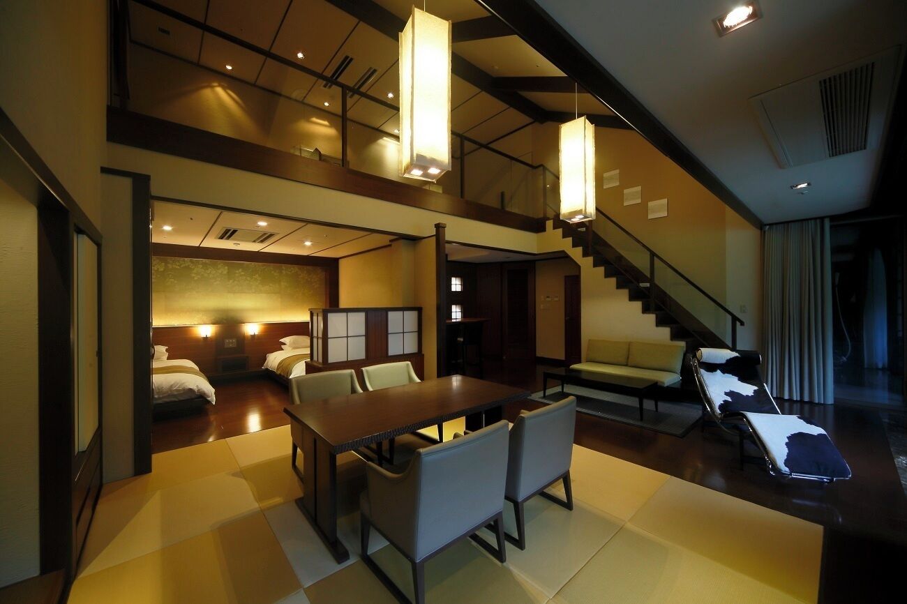A hotel room with a dining area, a sofa, a chair, and a staircase.