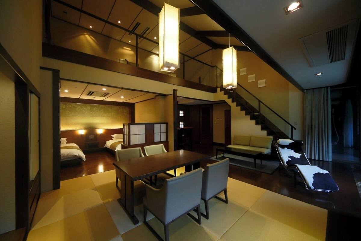 A hotel room with a dining area, a sofa, a chair, and a staircase.