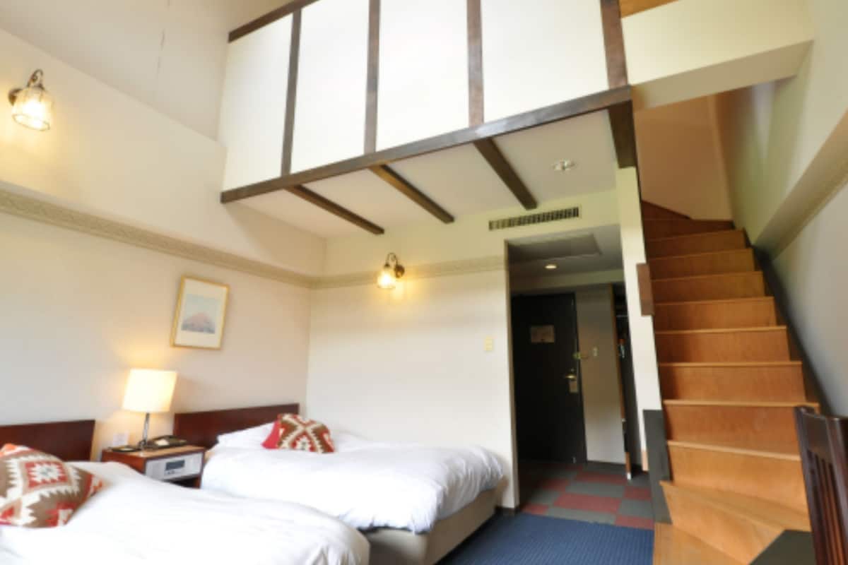Habitación de hotel con dos camas, una mesita de noche, una lámpara, un cuadro enmarcado en la pared y una escalera que lleva a otro nivel.