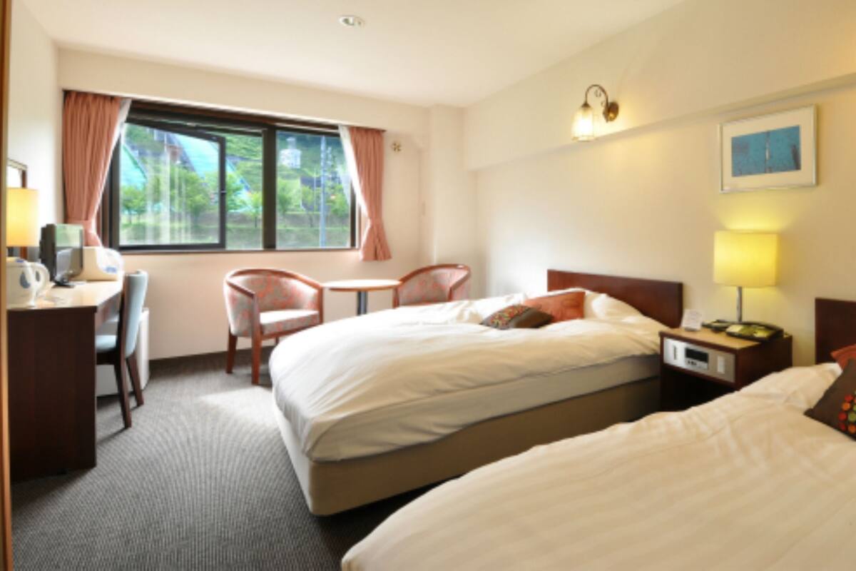 Ein Hotelzimmer mit zwei Betten, einem Schreibtisch, einem Stuhl, einem fenster mit Vorhängen und einem Nachttisch mit Lampe.