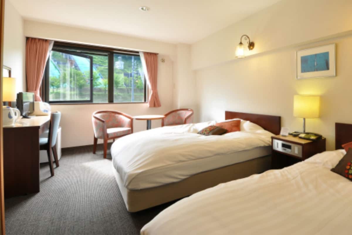 Habitación de hotel con dos camas, un escritorio, una silla, una ventana con cortinas y una mesita de noche con lámpara.