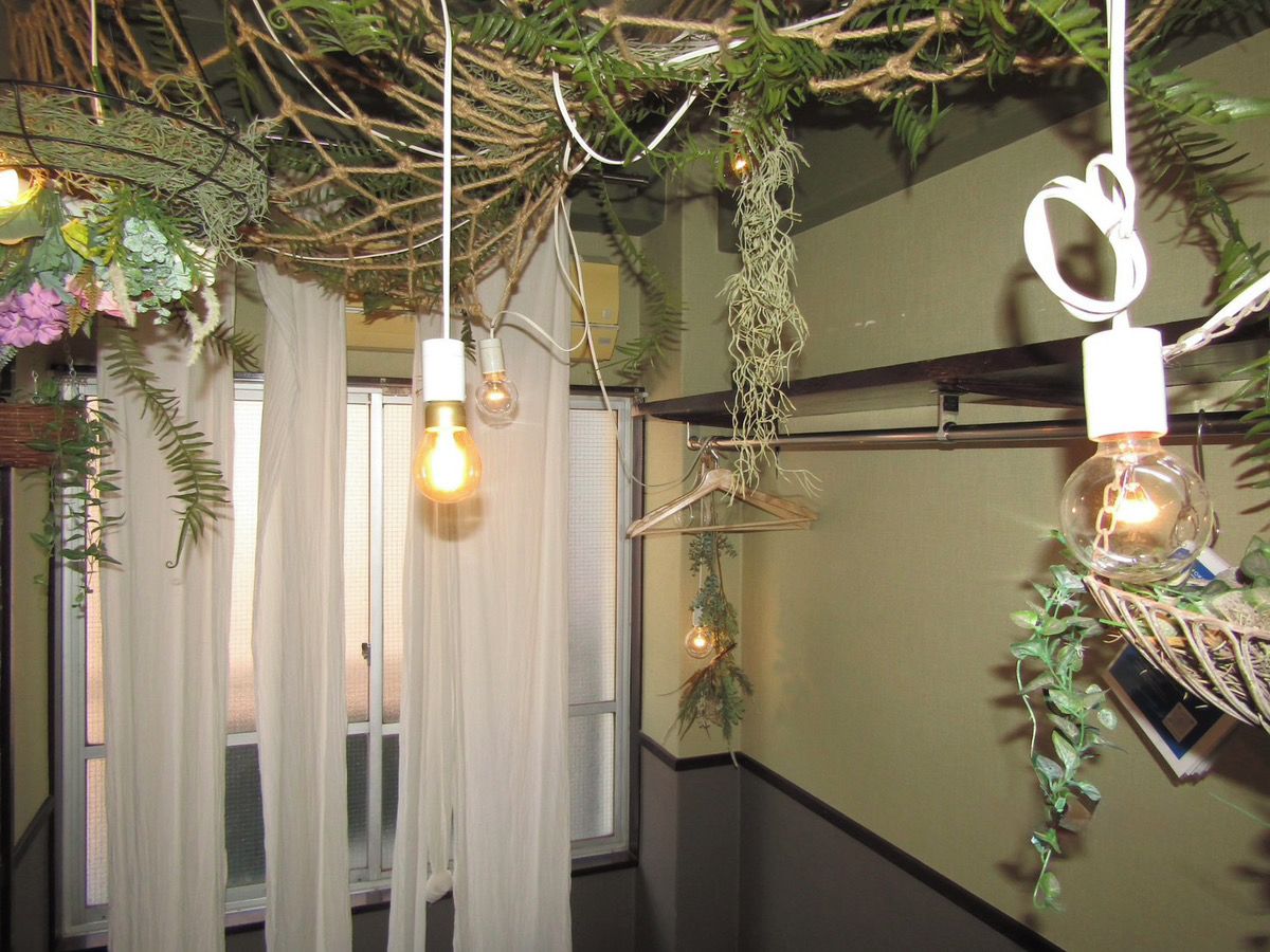 A room with a window covered by sheer curtains, a hanging light fixture, and a wall-mounted shelf with decorative items.