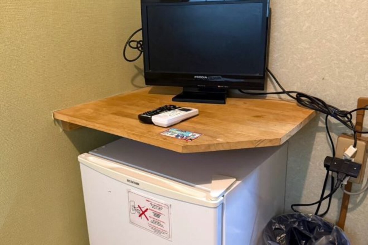 A television on a wooden desk with a remote control and a small box, a refrigerator with a warning label, and a trash bin.
