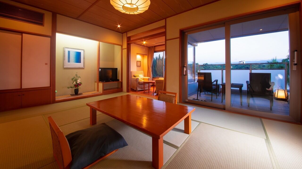 A traditional Japanese-style room with tatami flooring, a wooden dining table, a television, and a sliding glass door leading to an outdoor area with a view of the landscape.