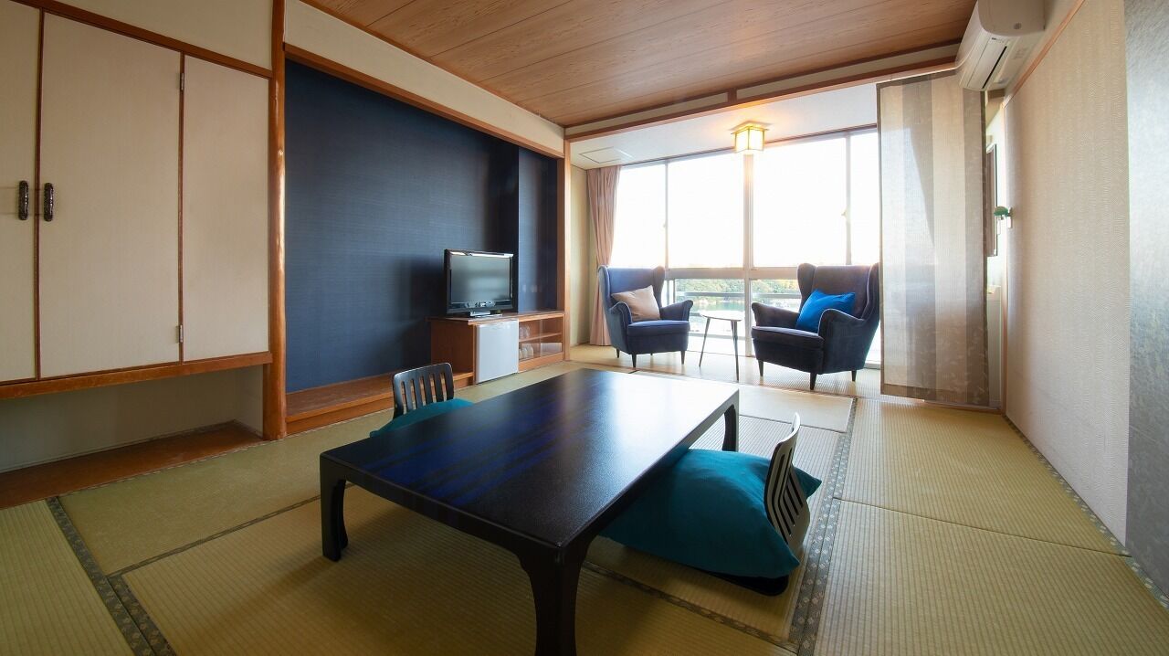 A traditional Japanese room with tatami flooring, a low table, chairs, a television, and a sliding door leading to a balcony with a view.