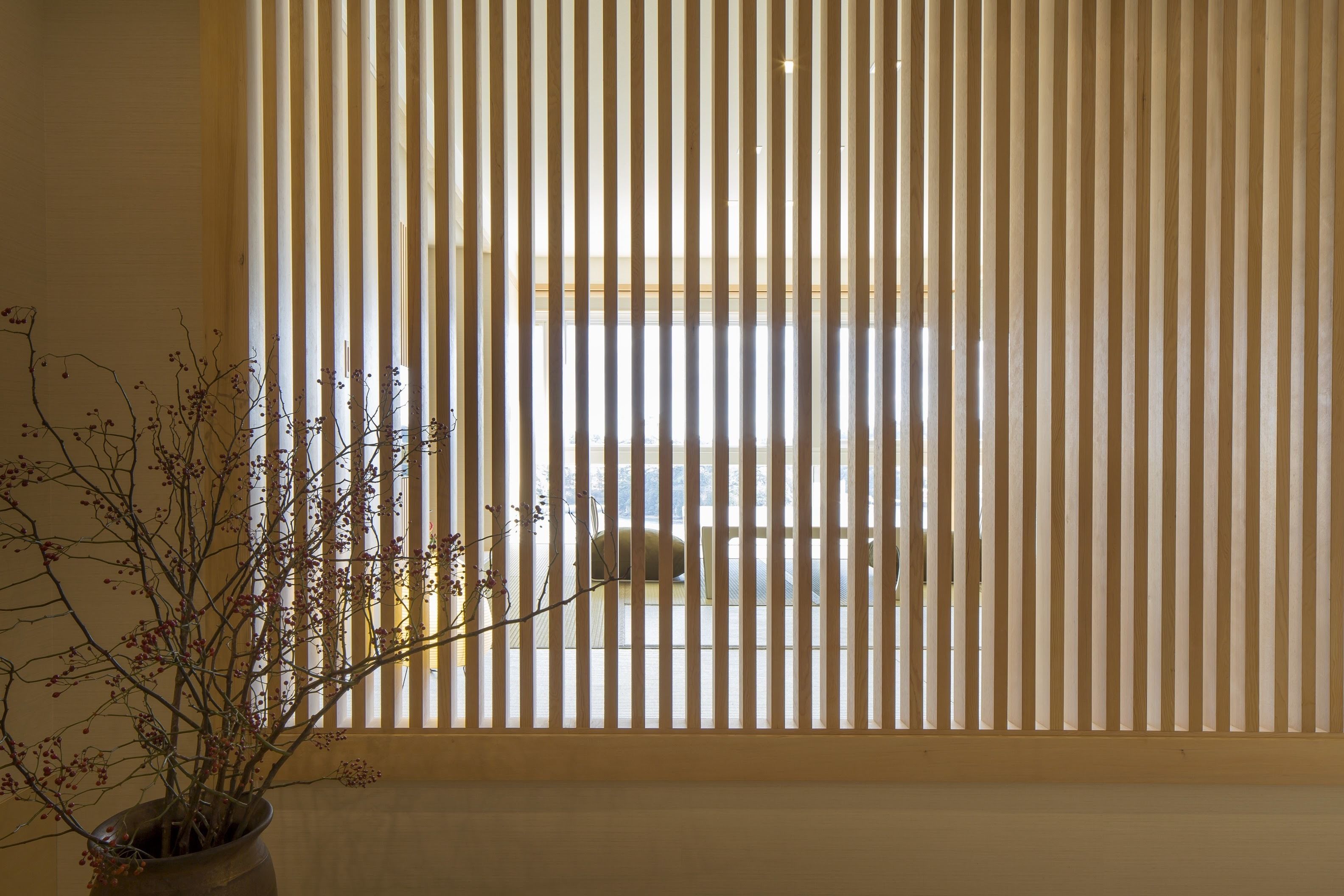 A room with vertical blinds, a potted plant, and a glimpse of a bed and window in the background.
