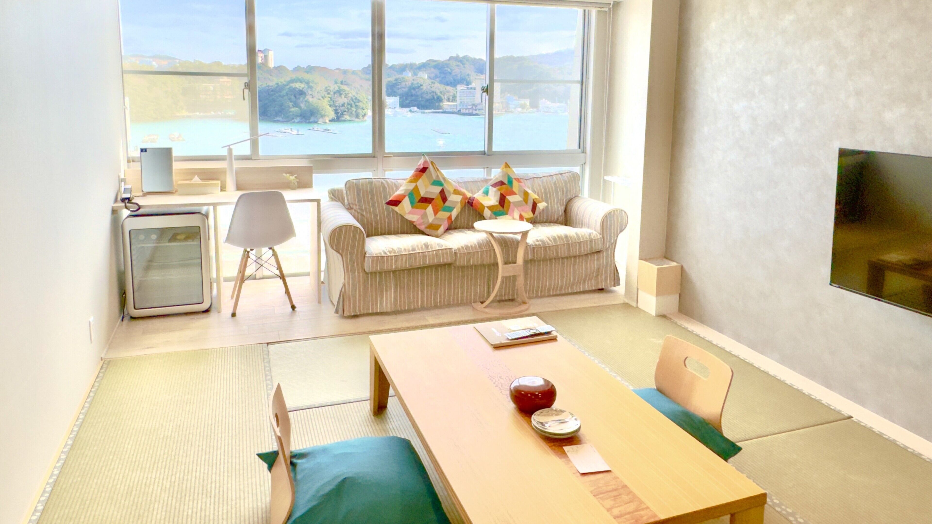A modern living room with a sofa, a small table, a TV, and a view of a body of water through large windows.