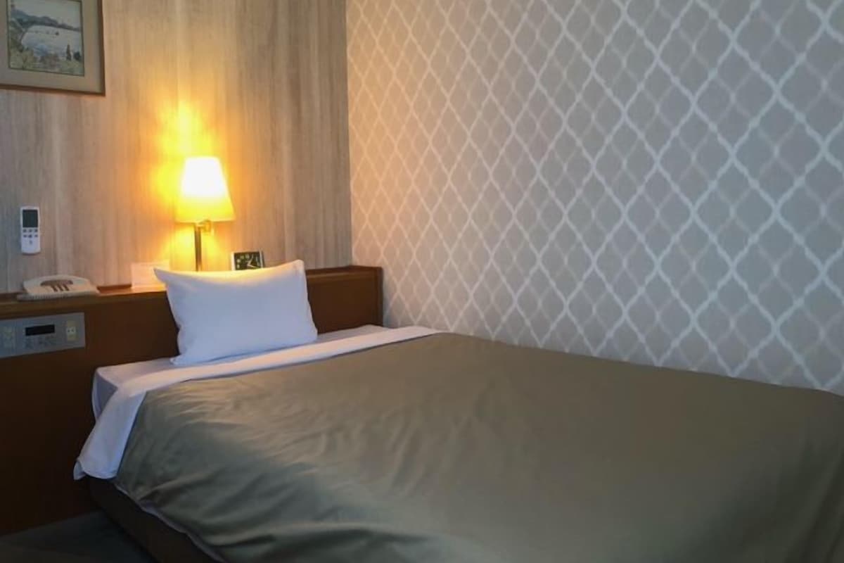 A hotel room with a bed, a nightstand with a lamp, a clock, and a phone.