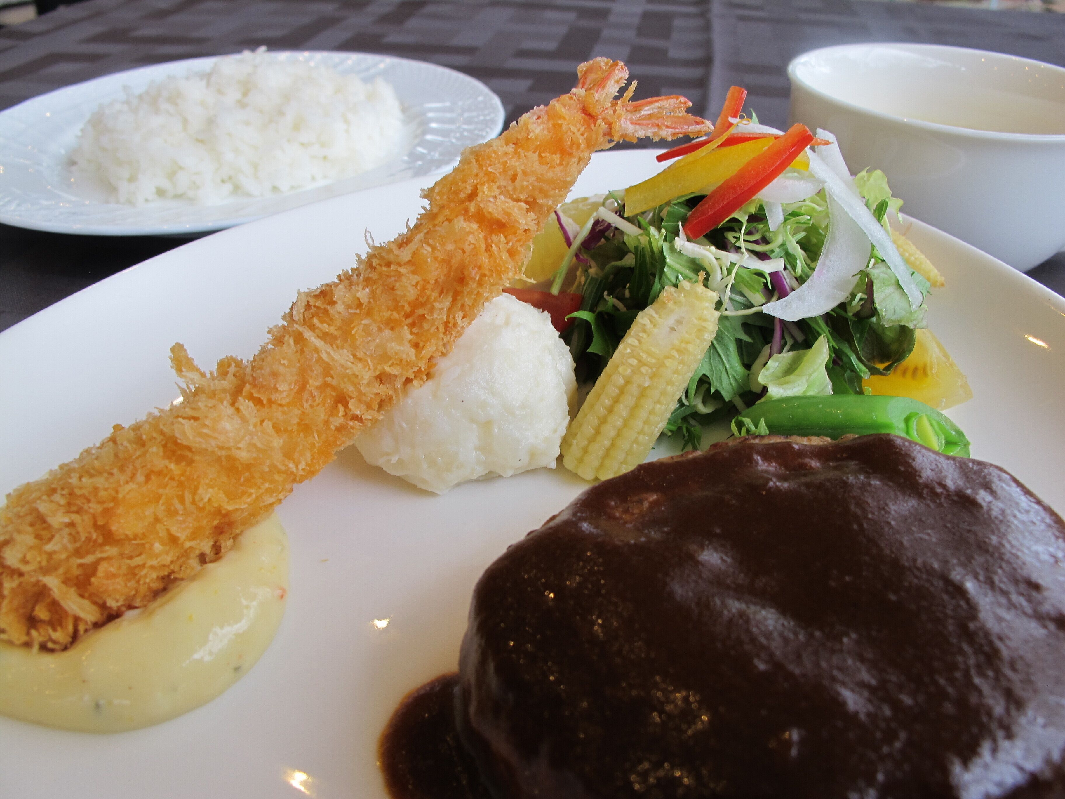 A plate with rice, a fried shrimp, a mound of white food, a green vegetable, and a dark sauce.