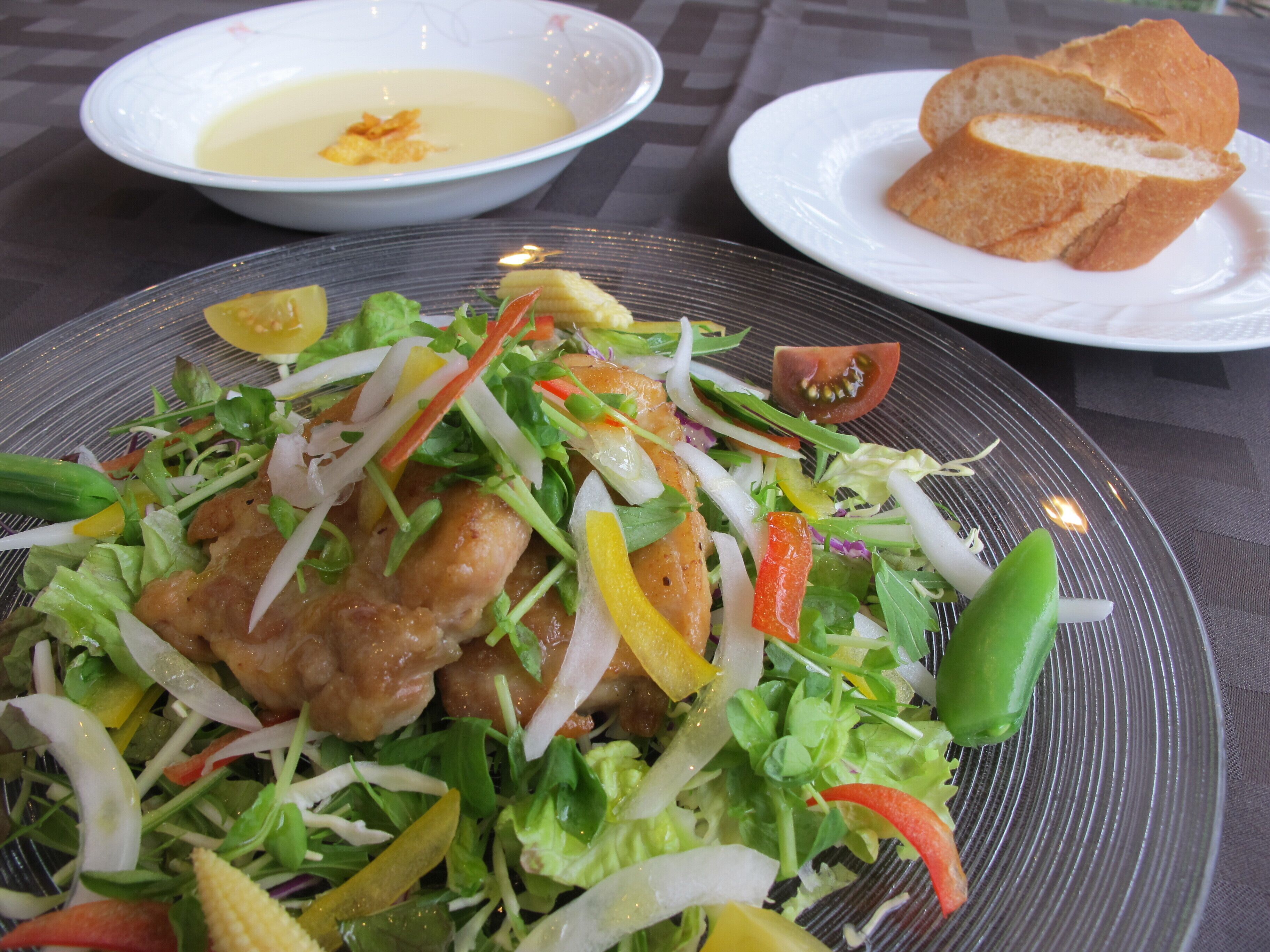 Un plato de ensalada con diversas verduras y un trozo de pollo, acompañado de un tazón de sopa y una rebanada de pan.