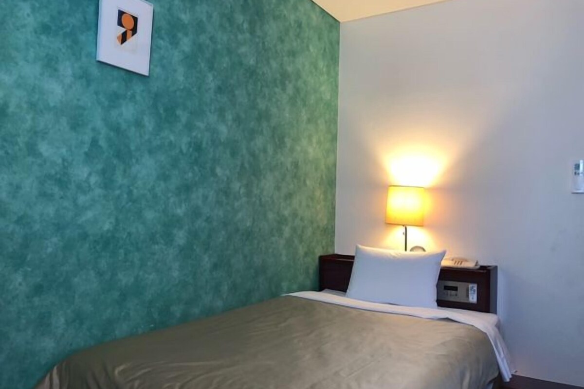 Un lit simple dans une chambre aux murs de couleur turquoise et au plafond blanc. Il y a une table de chevet avec une lampe et une petite horloge.
