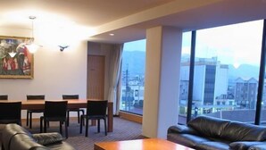 Une chambre d’hôtel dotée d’un canapé en cuir, d’une table basse en bois, d’un coin repas avec une table et des chaises, et de grandes fenêtres offrant une vue sur la ville.