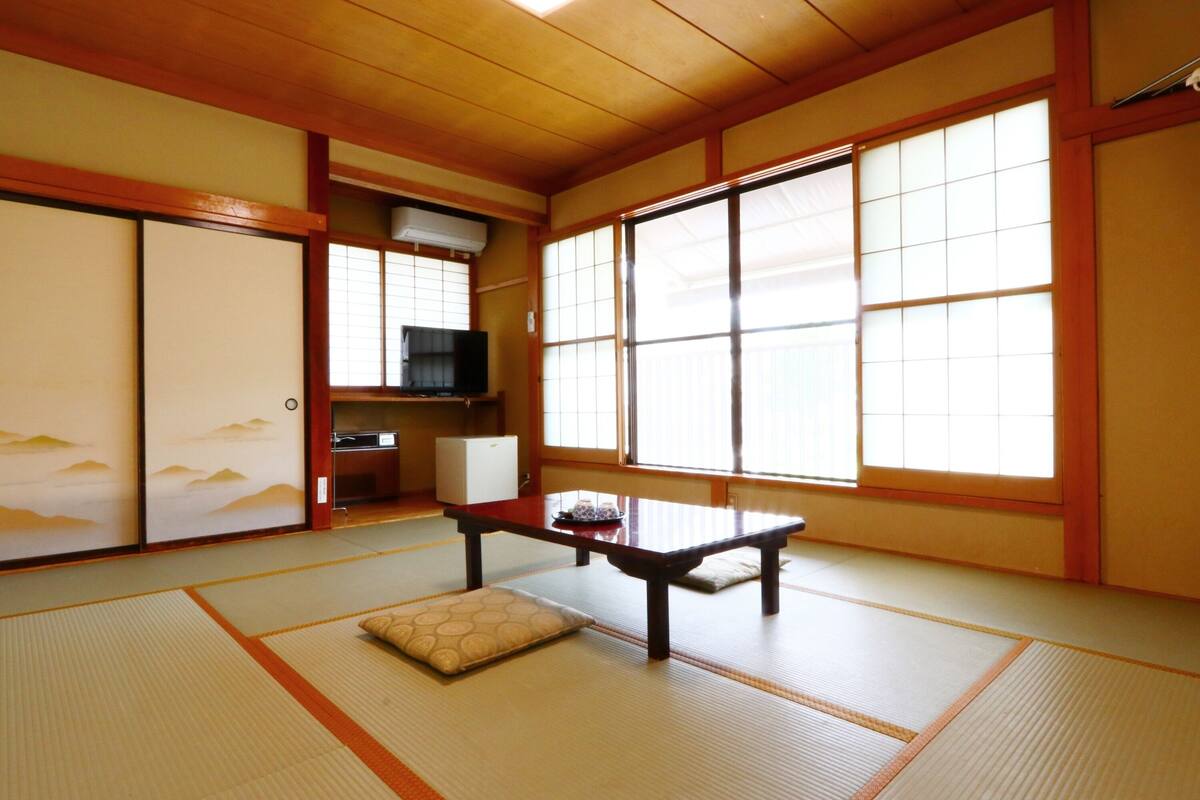 A traditional Japanese room with tatami flooring, a low table, a cushion, sliding doors, and a television.
