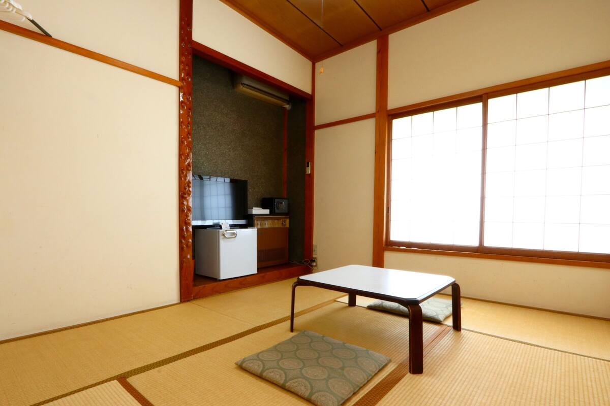 A traditional Japanese room with tatami flooring, a low table, a cushion, and a kitchenette area with a refrigerator and microwave.