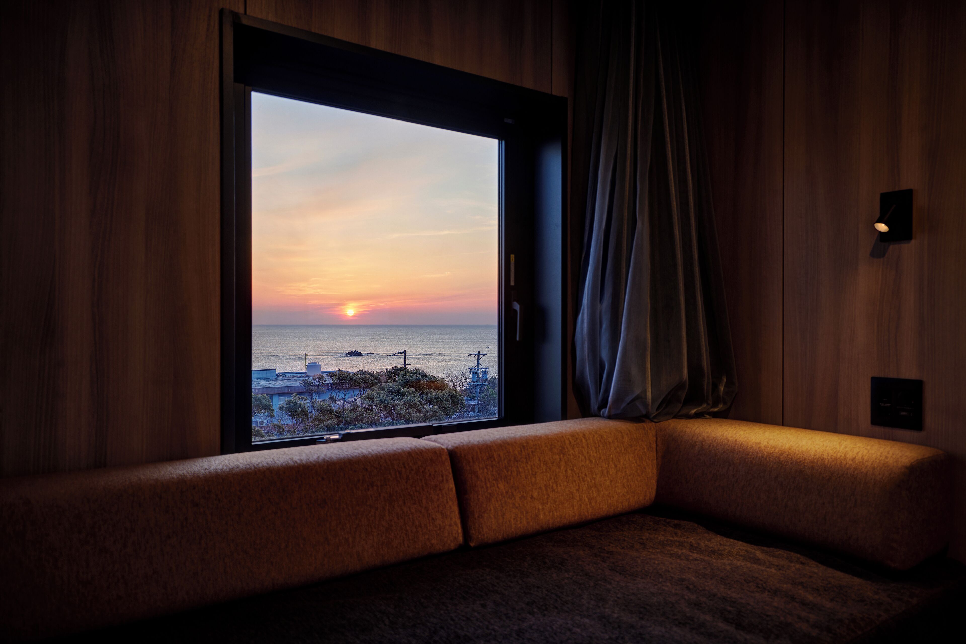 A room with a large window overlooking a sunset, a cushioned bench, and a wall-mounted light fixture.
