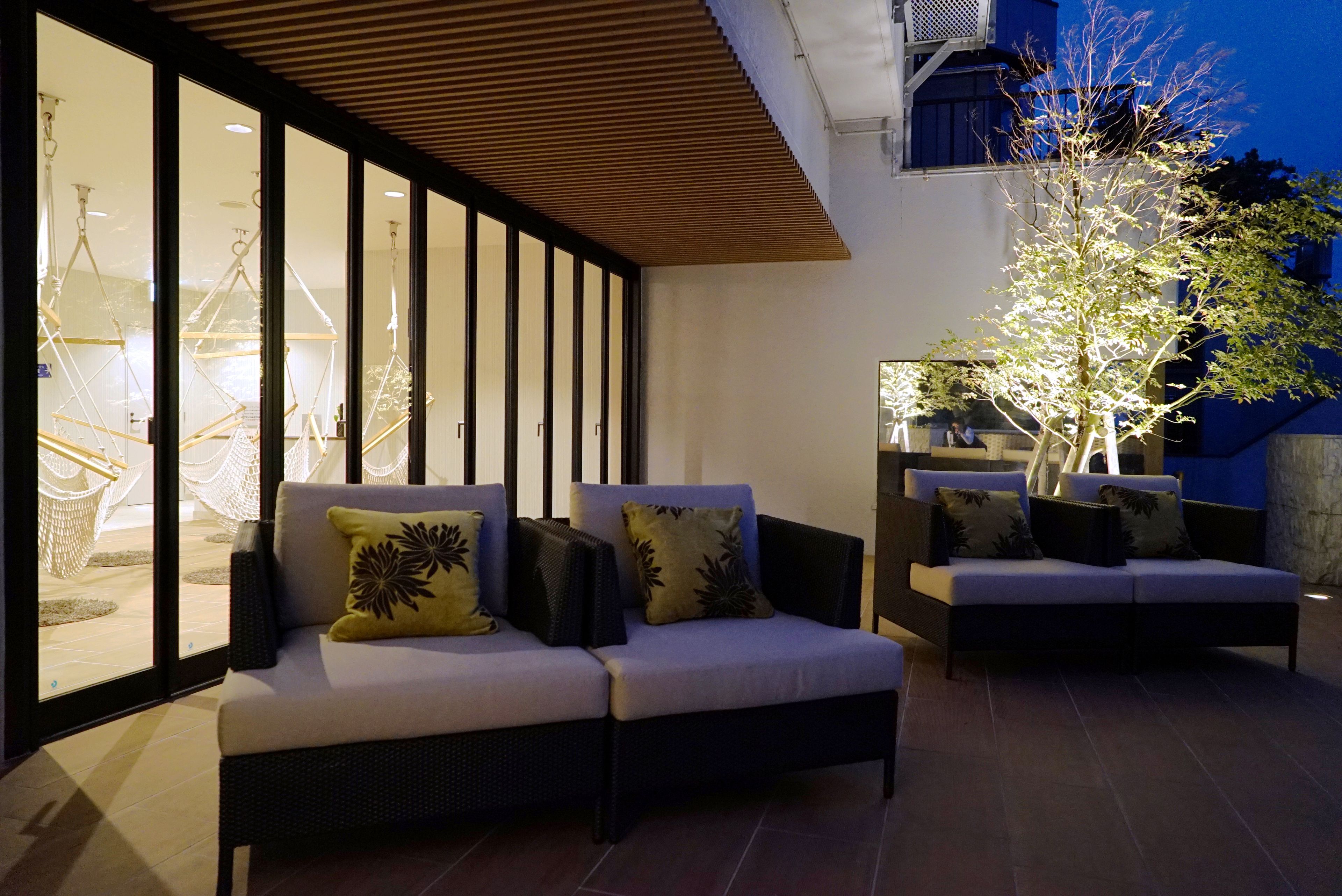 Un espace extérieur moderne aménagé avec deux canapés et une table à manger, entouré de grandes fenêtres et d’un arbre.