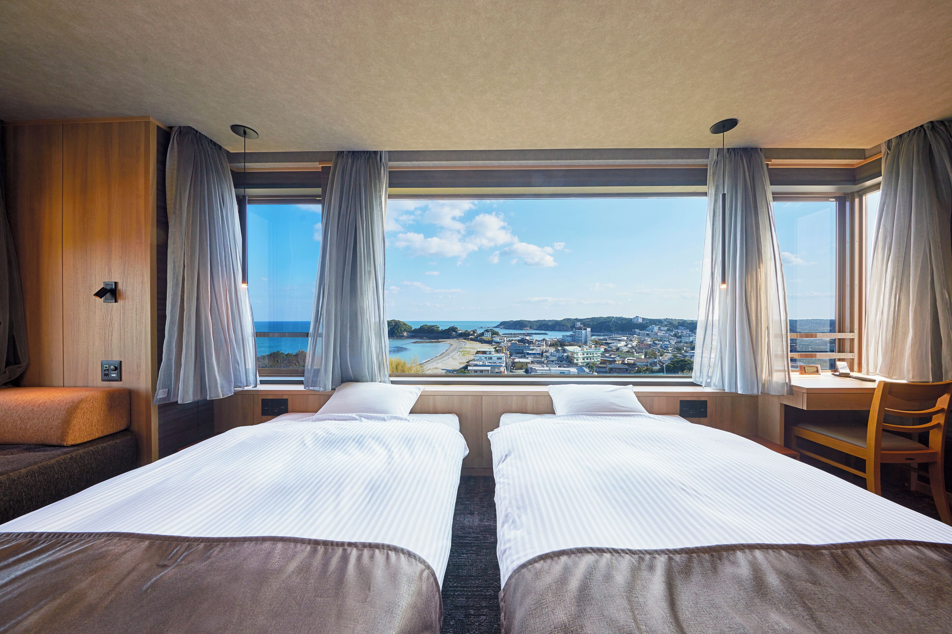 Ein Hotelzimmer mit zwei Betten, einem Schreibtisch und Blick auf eine Küstenstadt.
