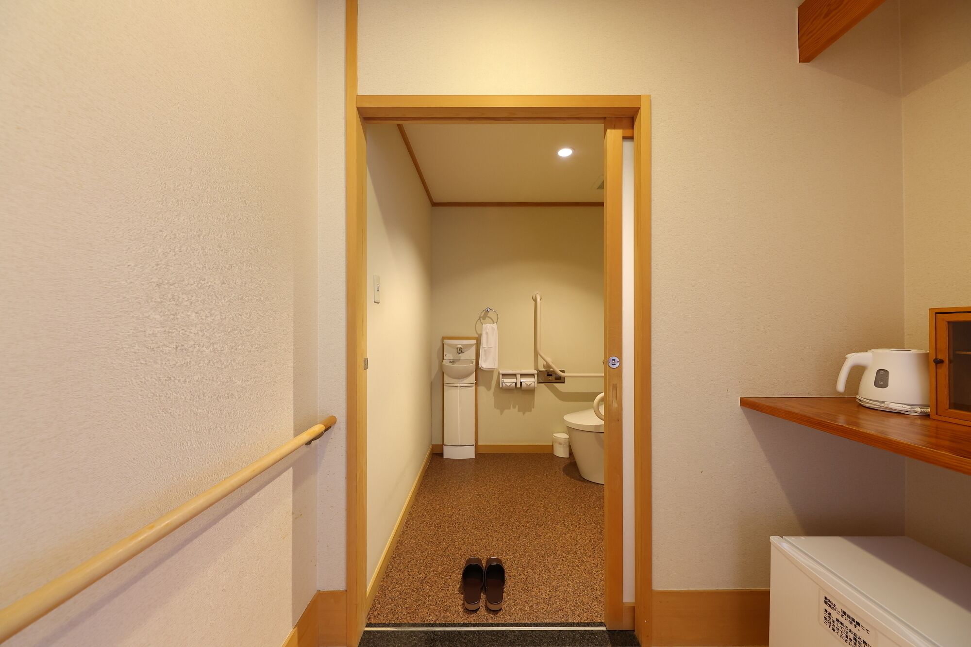 A narrow hallway leading to a bathroom with a toilet, sink, and shower. There is a wooden shelf with a white appliance on it, and a pair of shoes on the floor.