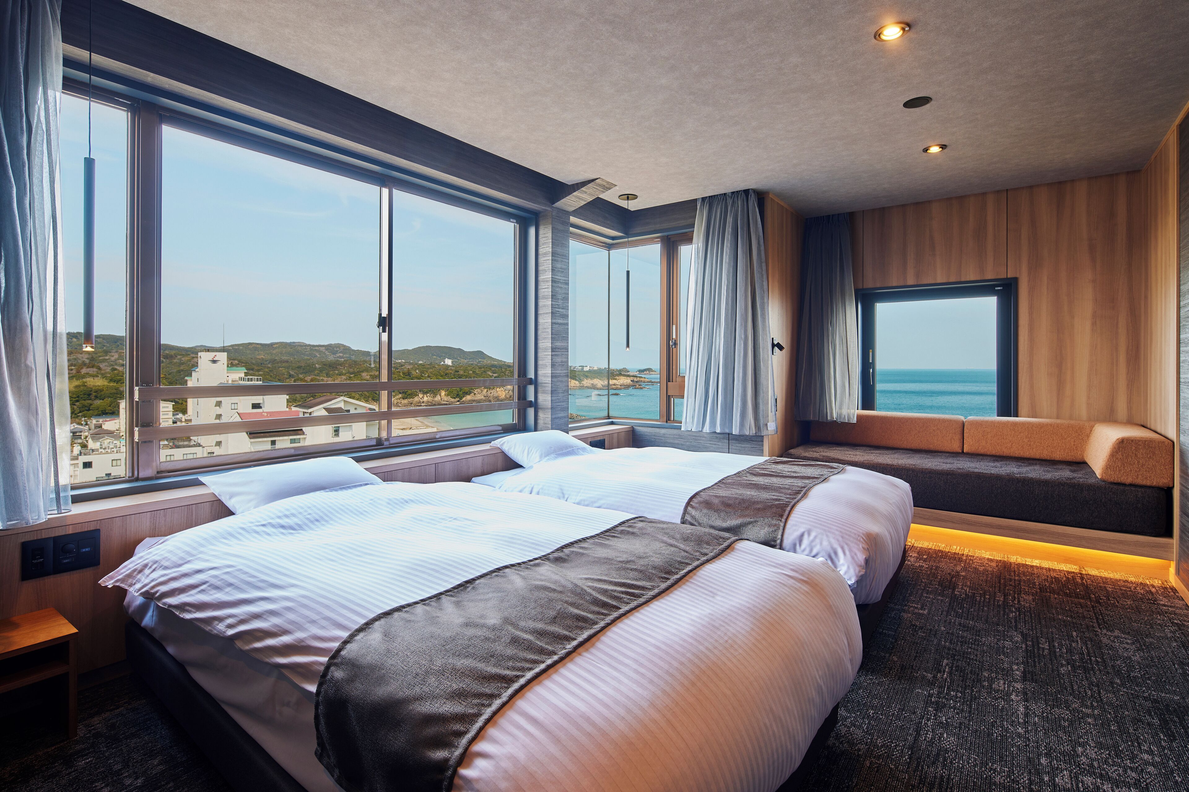 Ein Hotelzimmer mit zwei Betten, großen Fenstern, Meerblick und einer fest eingebauten Sitzbank.