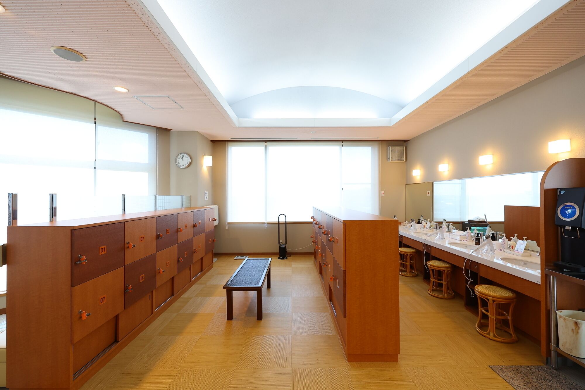 A modern hotel bathroom with wooden lockers, a bench, and a long counter with toiletries.