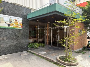 A hotel entrance with a glass door, a small tree in the foreground, and a signboard above the entrance.