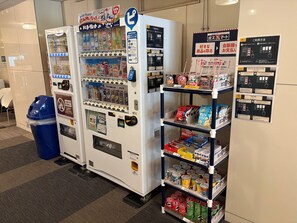 Automat mit Snacks und Getränken, eine blaue Mülltonne und ein Regal mit verpackten Waren.