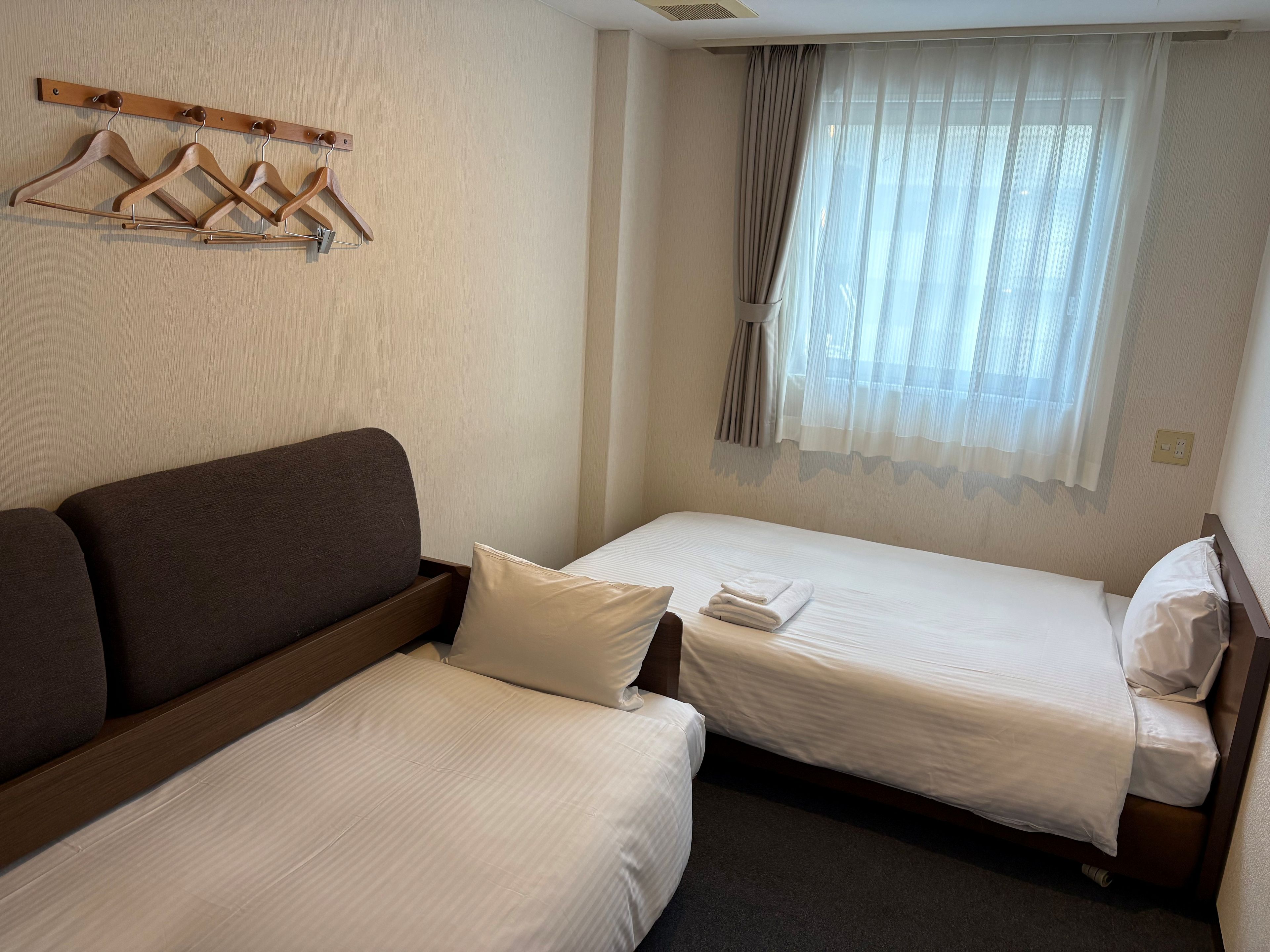 A hotel room with two single beds, a wooden sofa, a coat rack with hangers, and a window with sheer curtains.