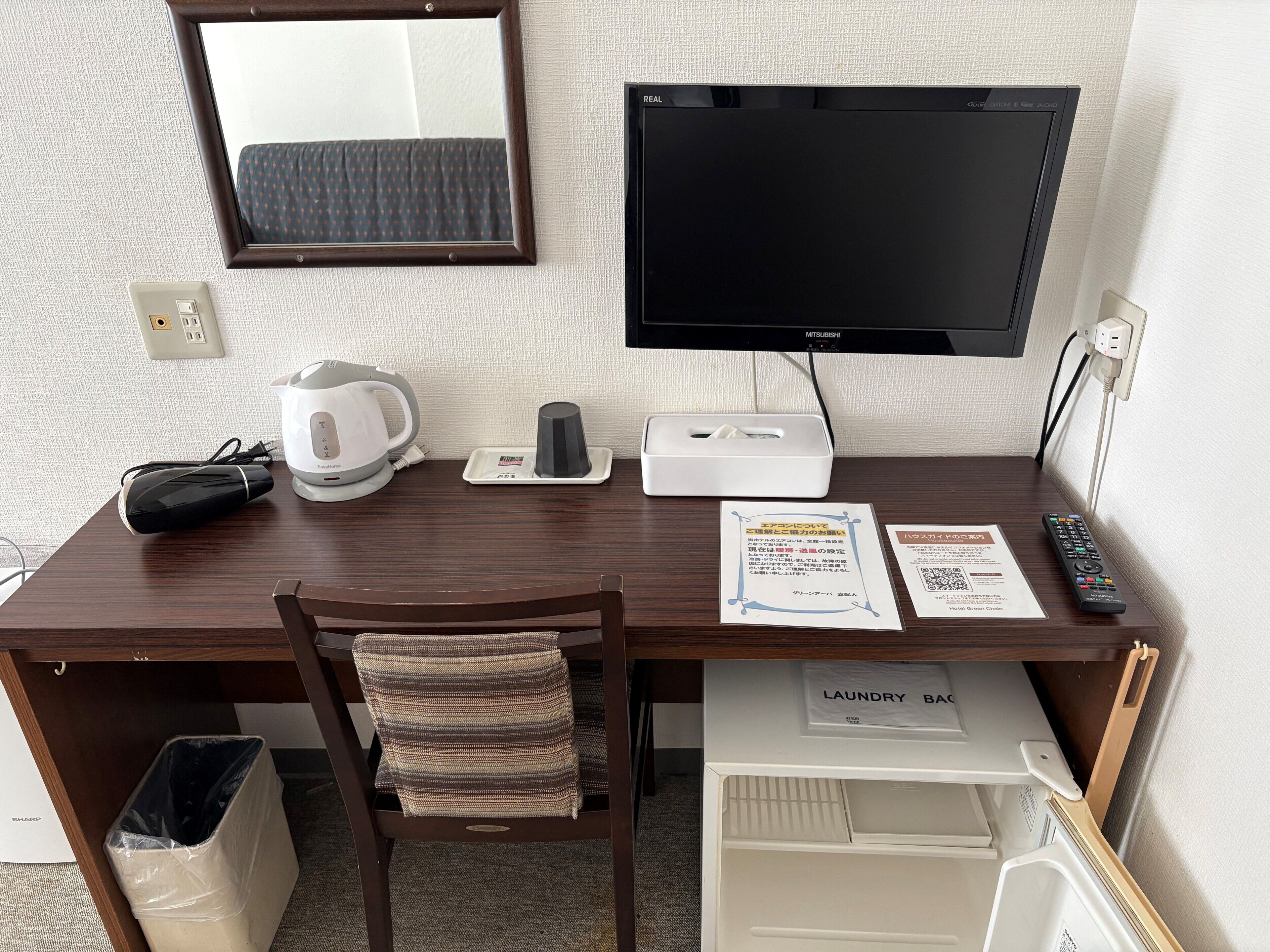 Une chambre d’hôtel avec un bureau en bois, un écran noir, une bouilloire, une télécommande, une poubelle et un panier à linge.
