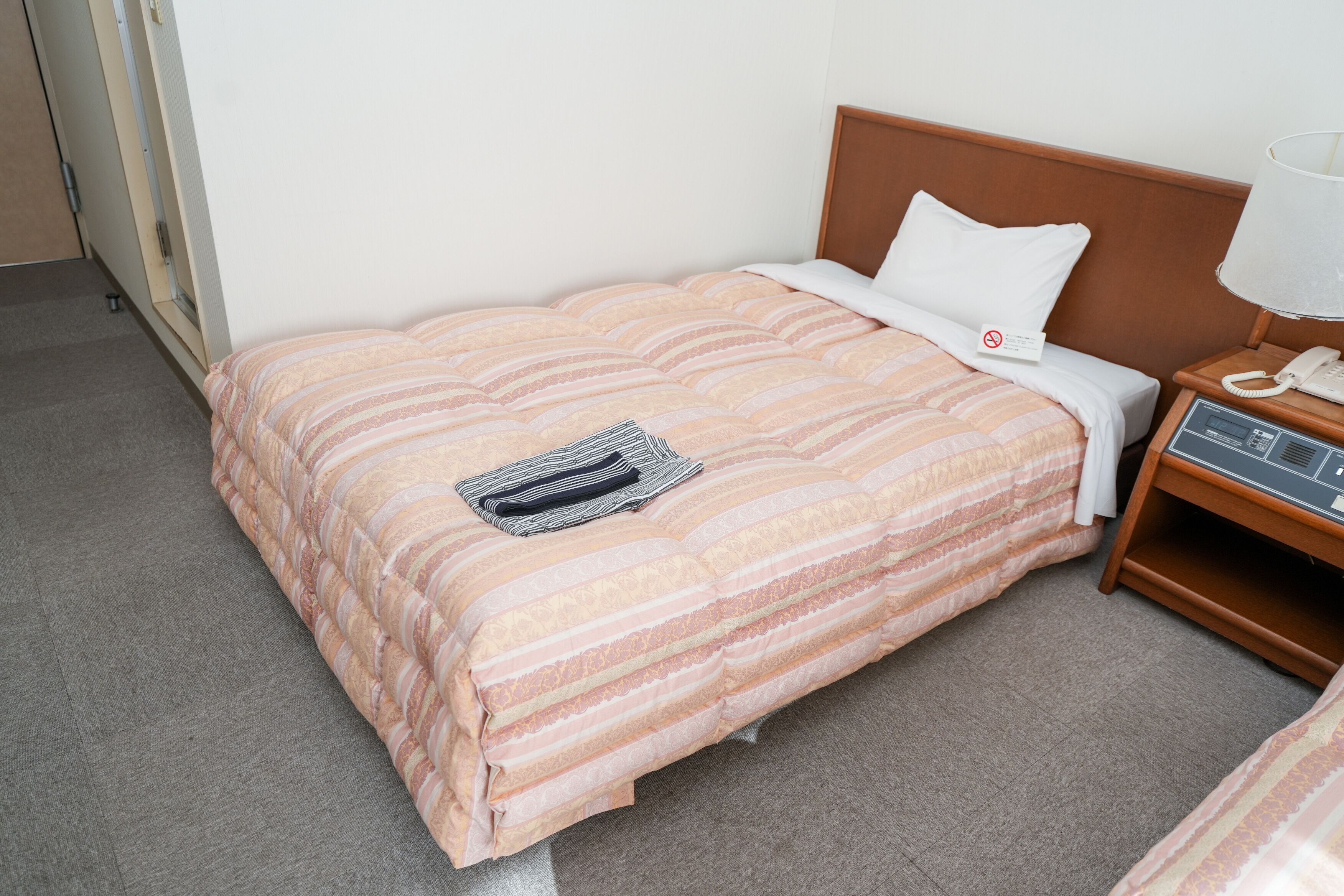 A single bed with a striped bedspread, a pillow, a bedside telephone, and a lamp on a wooden nightstand.