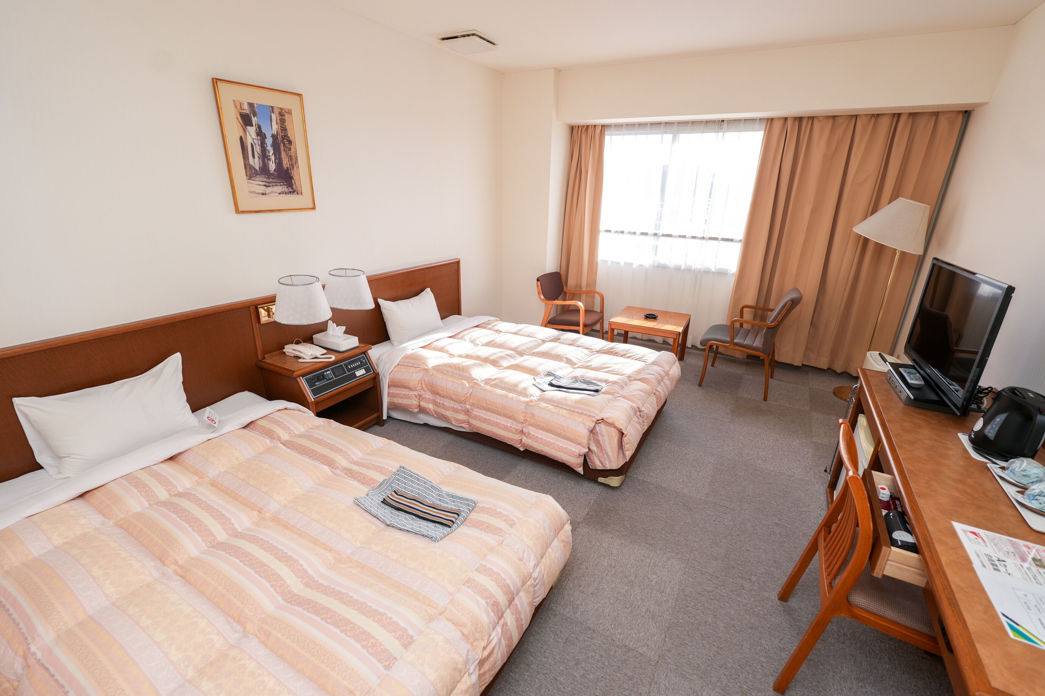 Ein Hotelzimmer mit zwei Betten, einem Schreibtisch, einem Fernseher und einem Fenster mit Vorhängen.