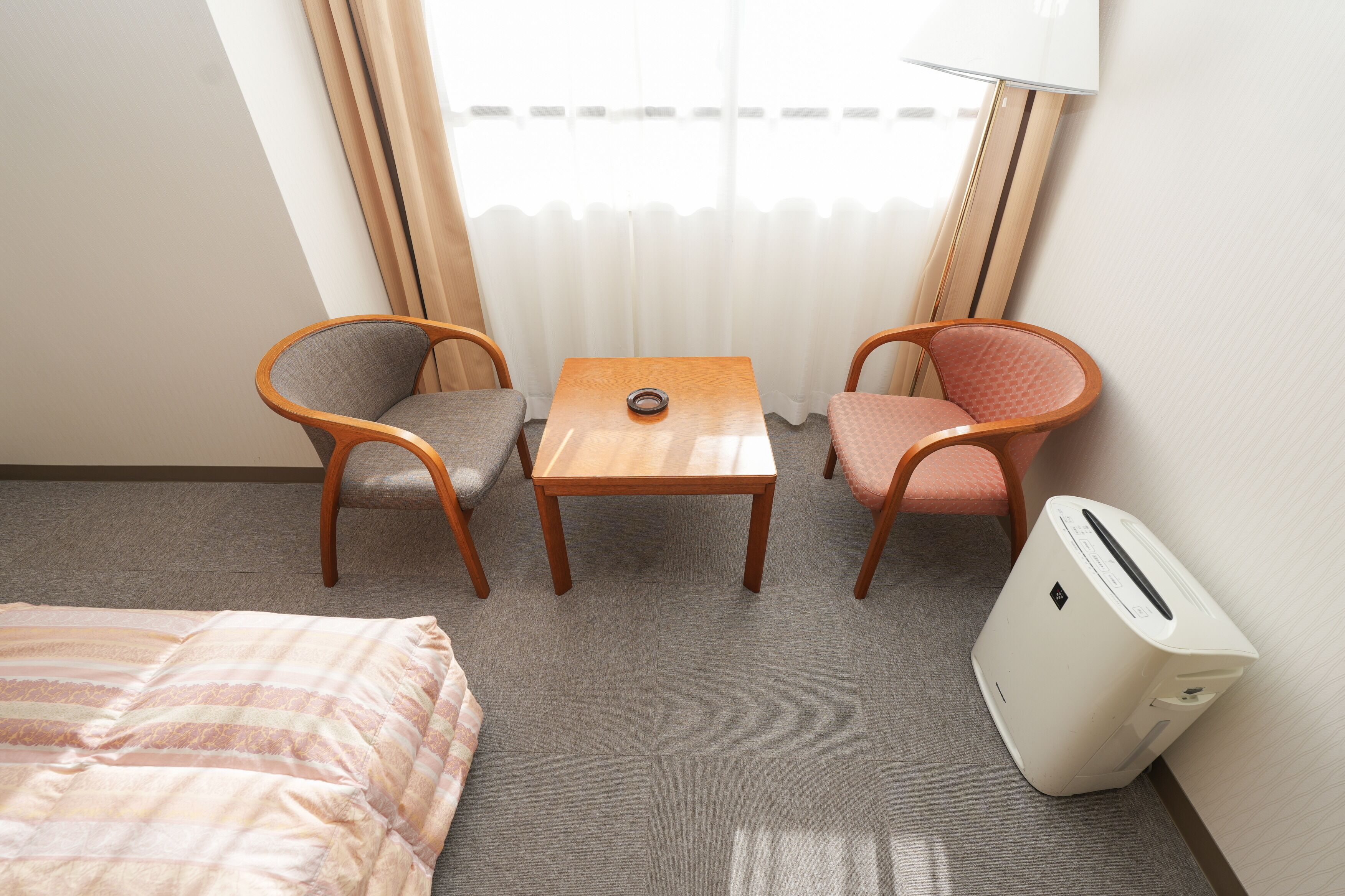 A hotel room with a bed, two chairs, a small table, a lamp, and a heater.