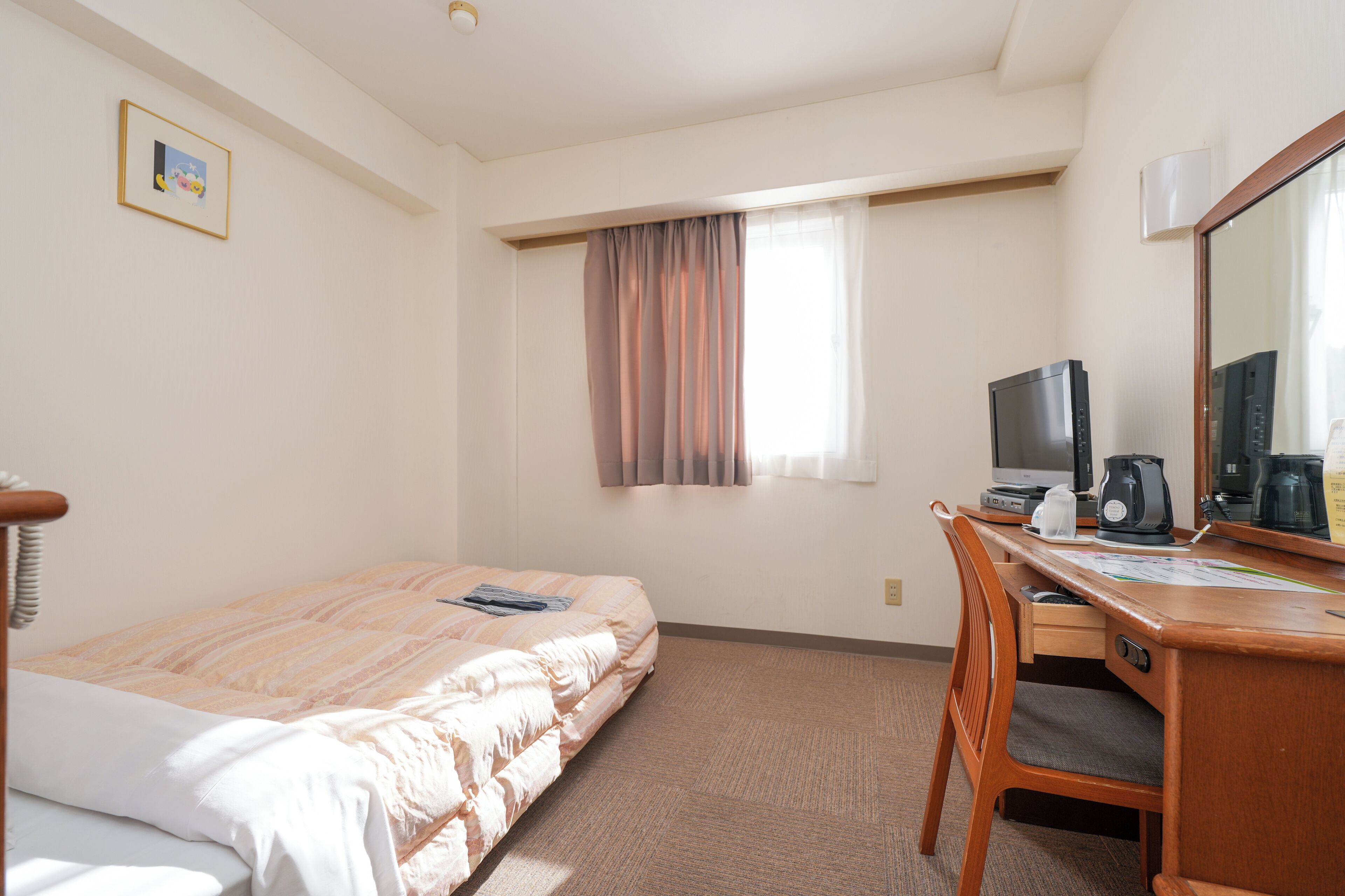 A hotel room with a bed, a desk with a computer, a chair, a window with curtains, and a picture on the wall.