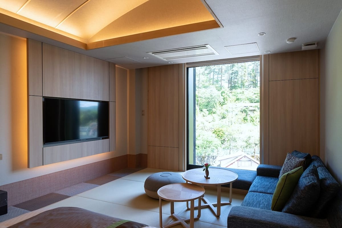 A modern living room with a flat-screen TV, a sofa, a small round table, tatami mats, and a large window with a view of greenery.