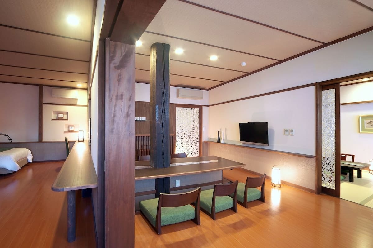 Una habitación de hotel con piso de madera, una cama, un escritorio, sillas, un televisor y un separador decorativo.