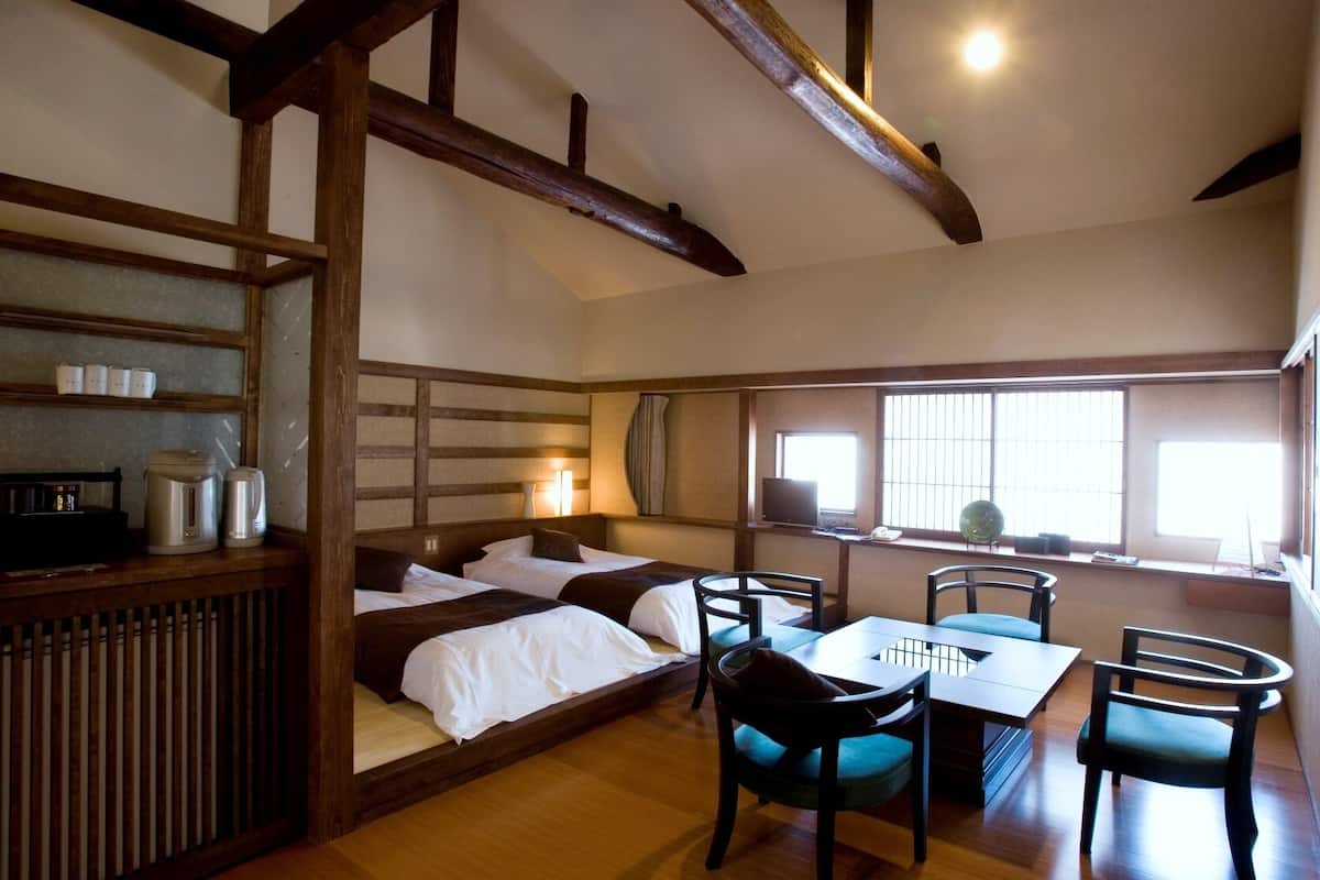 A traditional Japanese-style room with a low ceiling, wooden beams, a bed with white bedding, a small table with chairs, and a window with blinds.