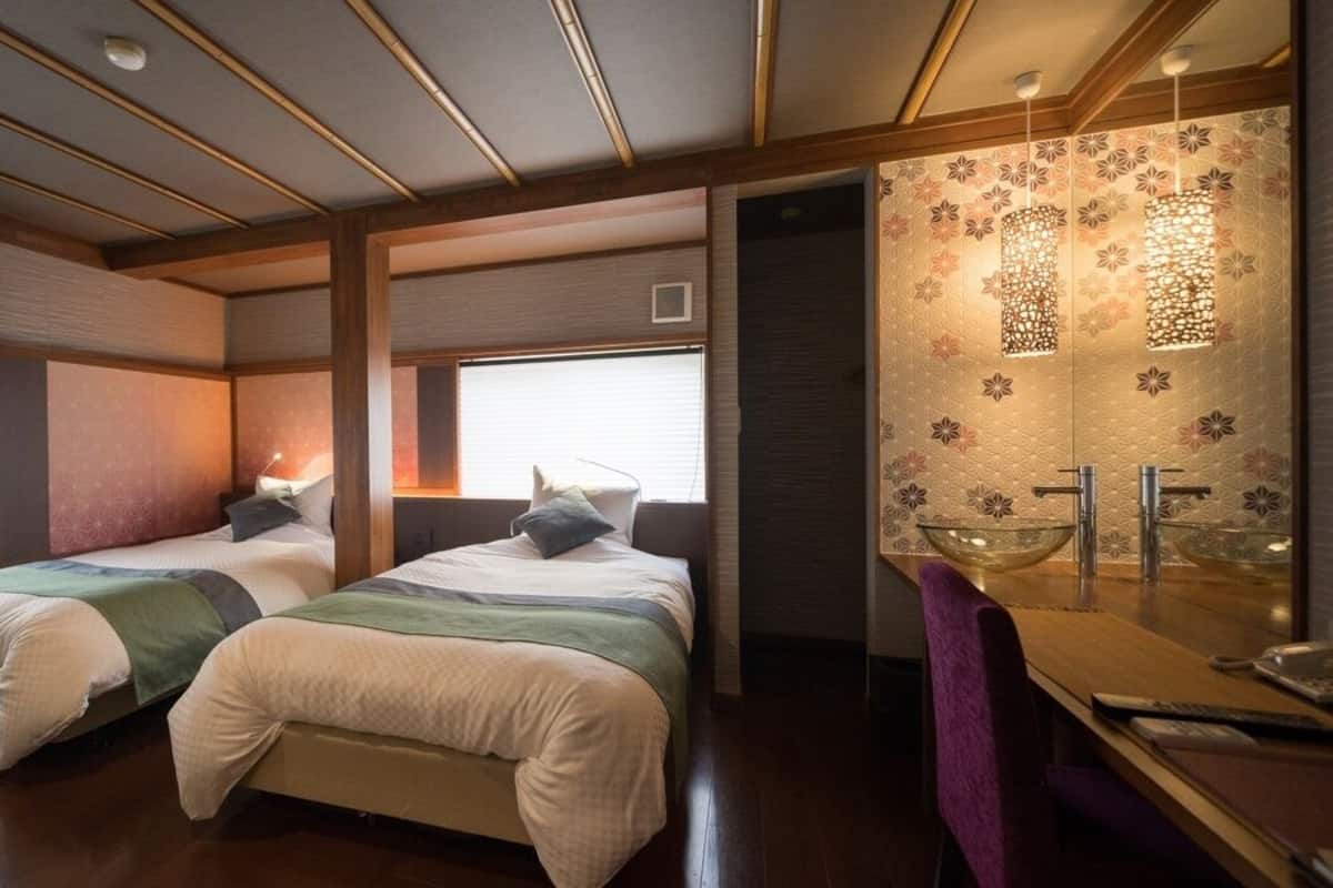 Habitación de hotel con dos camas, un escritorio y baño con espejo grande y azulejos decorativos.
