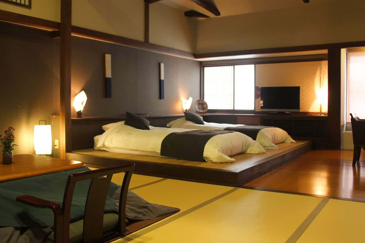 A traditional Japanese-style room with tatami flooring, a low bed with pillows, a television, and a wooden table with chairs.