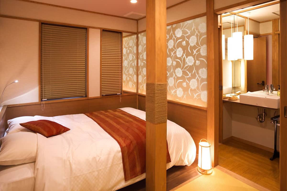 A room with a bed, a bedside lamp, a window with blinds, a wooden partition with floral wallpaper, and a bathroom with a sink and mirror.